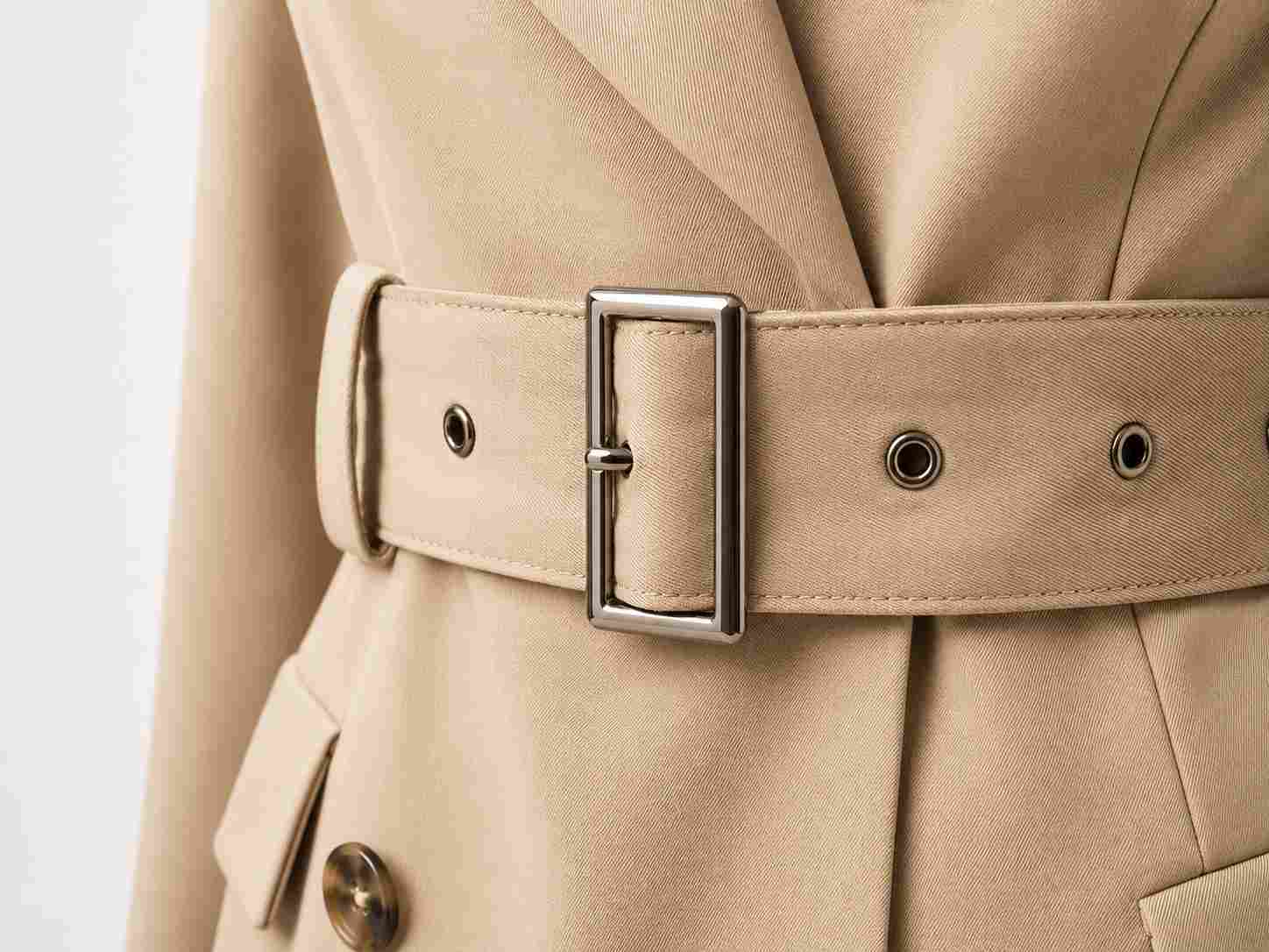 metal belt buckle, A silver metal belt buckle is attached to a wide beige fabric belt with metal eyelets on a beige garment.